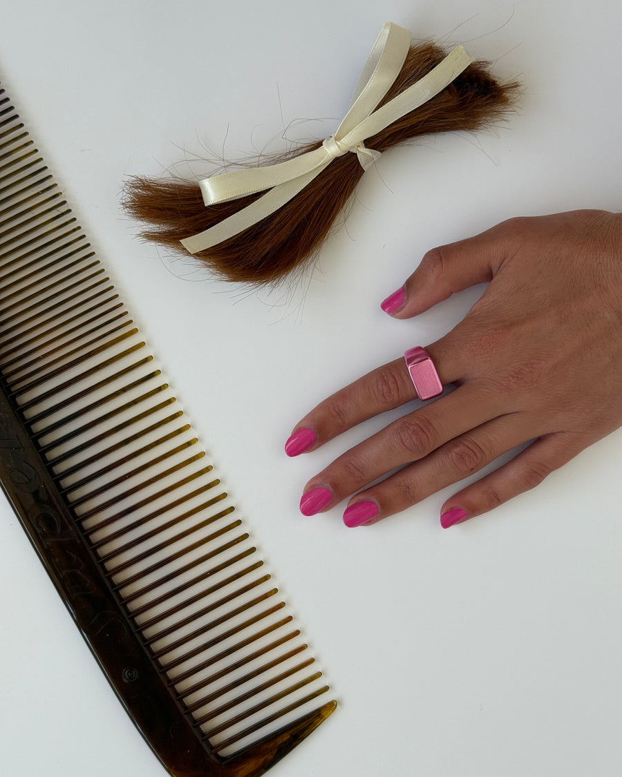 Hand with pink nails wearing the pink break in ring beside a comb and a cut ponytail with a bow