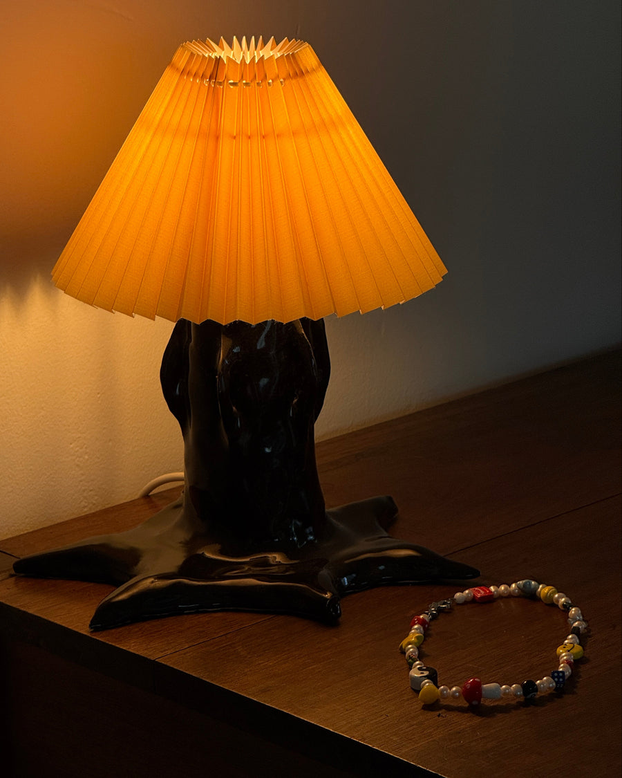 The gabe beaded charm necklace on a wooden table beside a hand-shaped lamp with an orange pleated shade