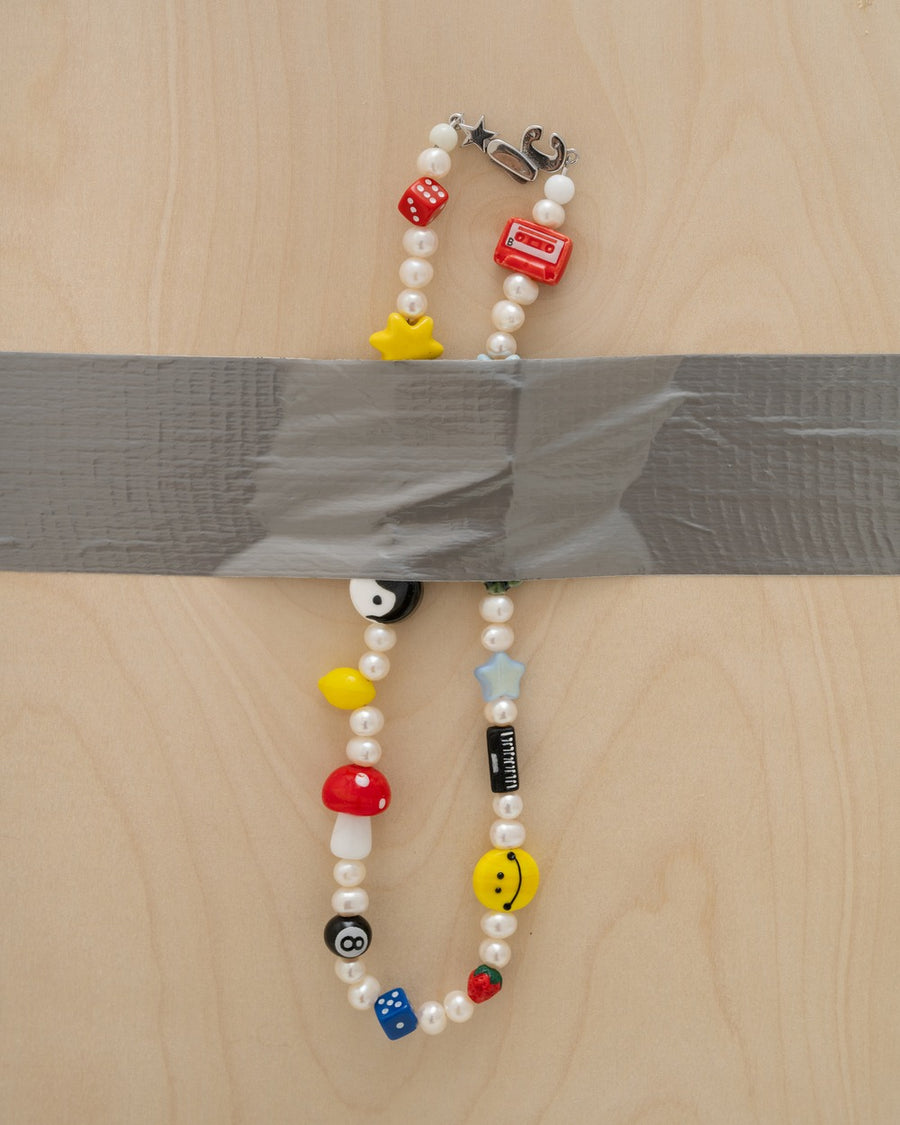 Pearl necklace with red mushroom, yellow smiley, star, yellow smiley face, lemon, yin-yang, casette, and dice beads taped to a plywood board – the gabe necklace