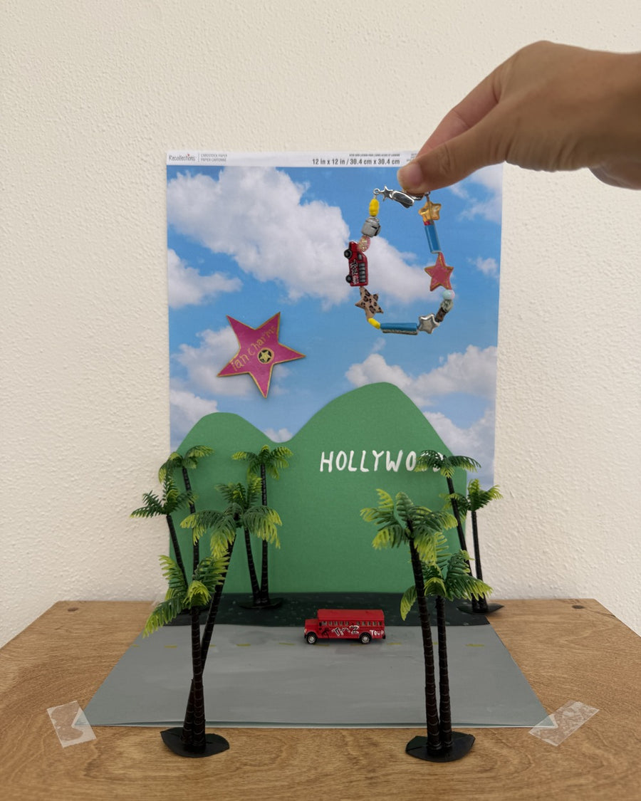 Hand holding the LA bracelet over a miniature LA hillside diorama with palm trees and a red bus