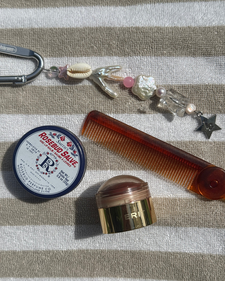 Marlee shell keychain flat lay with lip balm, a tortoiseshell comb and a gold compact on striped fabric