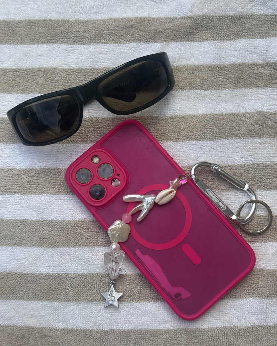 Pink phone case with the marlee shell keychain beside black sunglasses on a striped towel