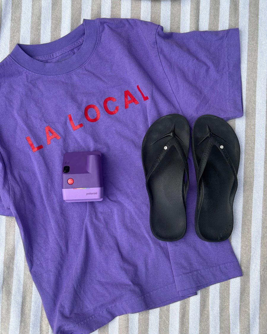 Flat lay of the purple la local tee with a purple instant camera and black flip-flops on a striped towel