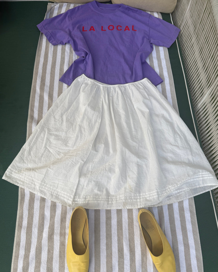 Purple la local tee and white skirt laid out on a striped beach towel with yellow flats