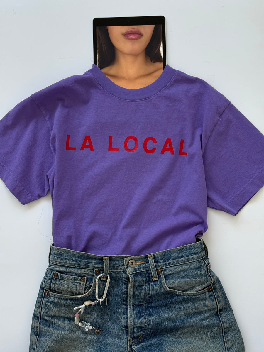 Person in the oversized purple la local tee with red lettering holding a mirror over their face