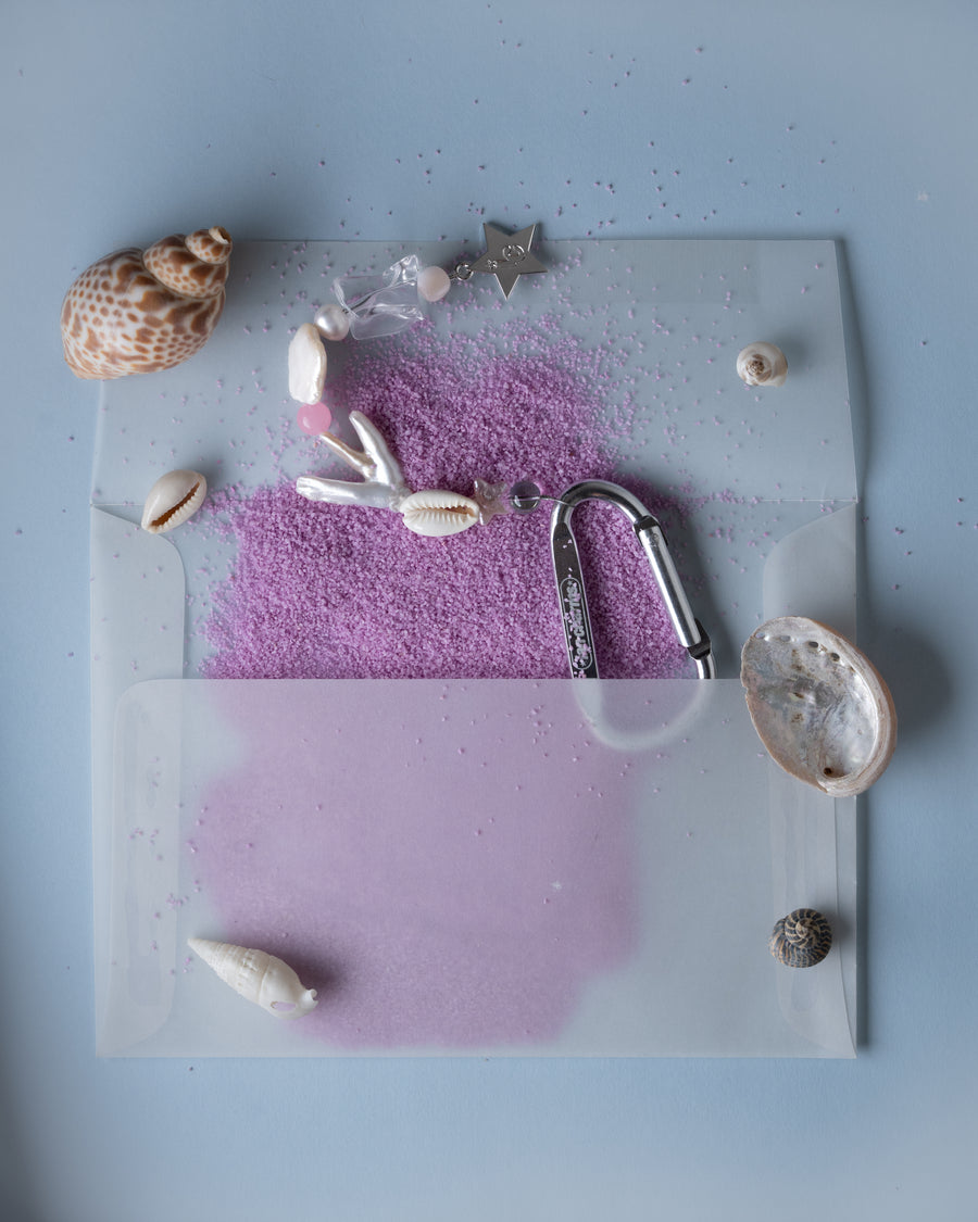 Marlee shell and pink bead keychain on a clear envelope with pink sand and seashells