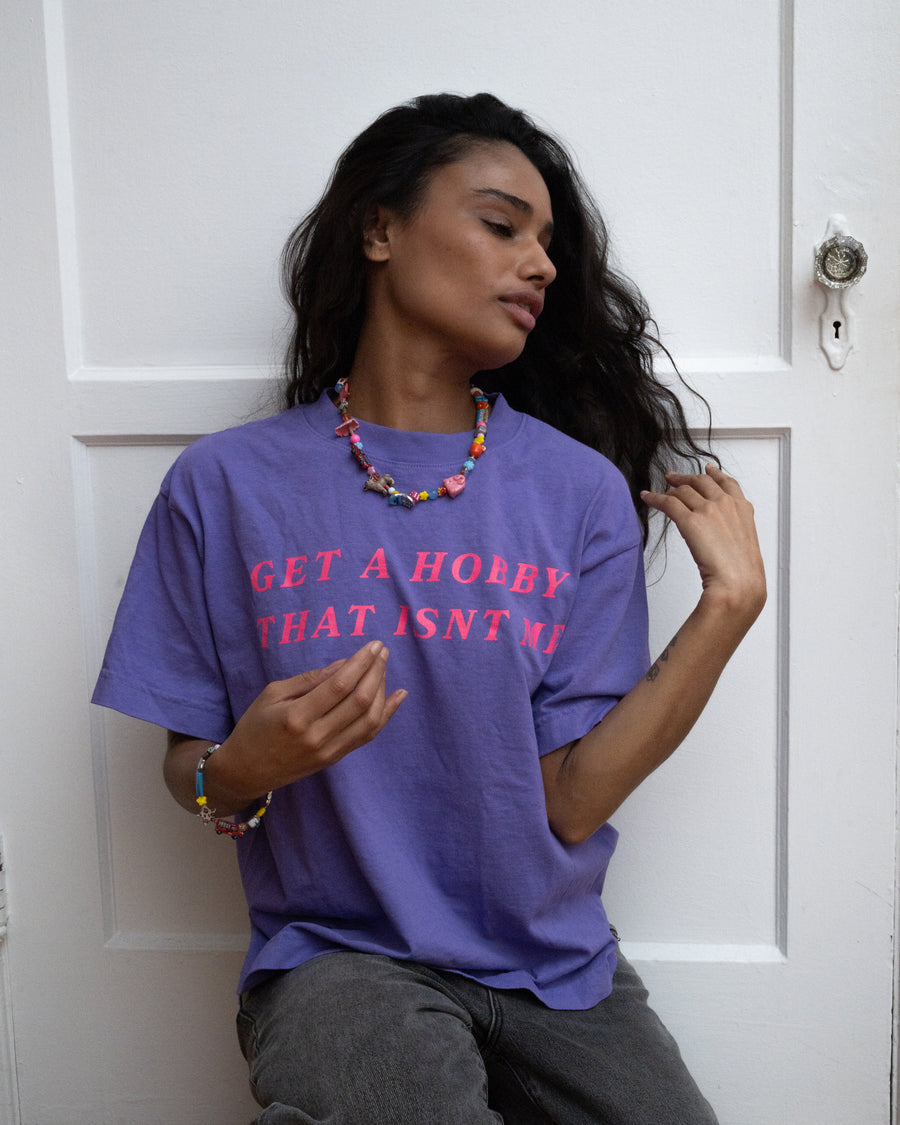 Woman sitting by a white door in the purple get a hobby tee and a beaded necklace