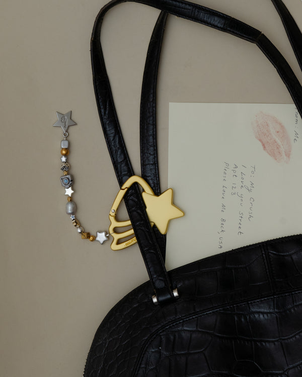 Gold shooting star carabiner keychain with gold and silver beads clipped to a black croc bag beside a lipstick-kissed letter – the good as gold keychain