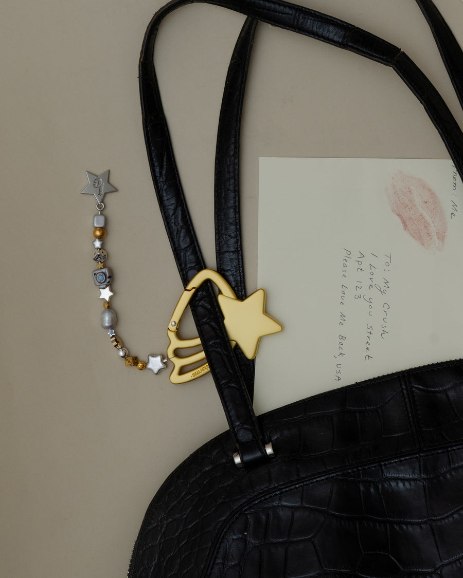 Gold shooting star carabiner keychain with gold and silver beads clipped to a black croc bag beside a lipstick-kissed letter – the good as gold keychain