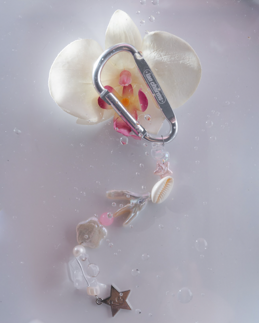 Silver engraved carabiner with shell and pink bead charms in front of a white orchid