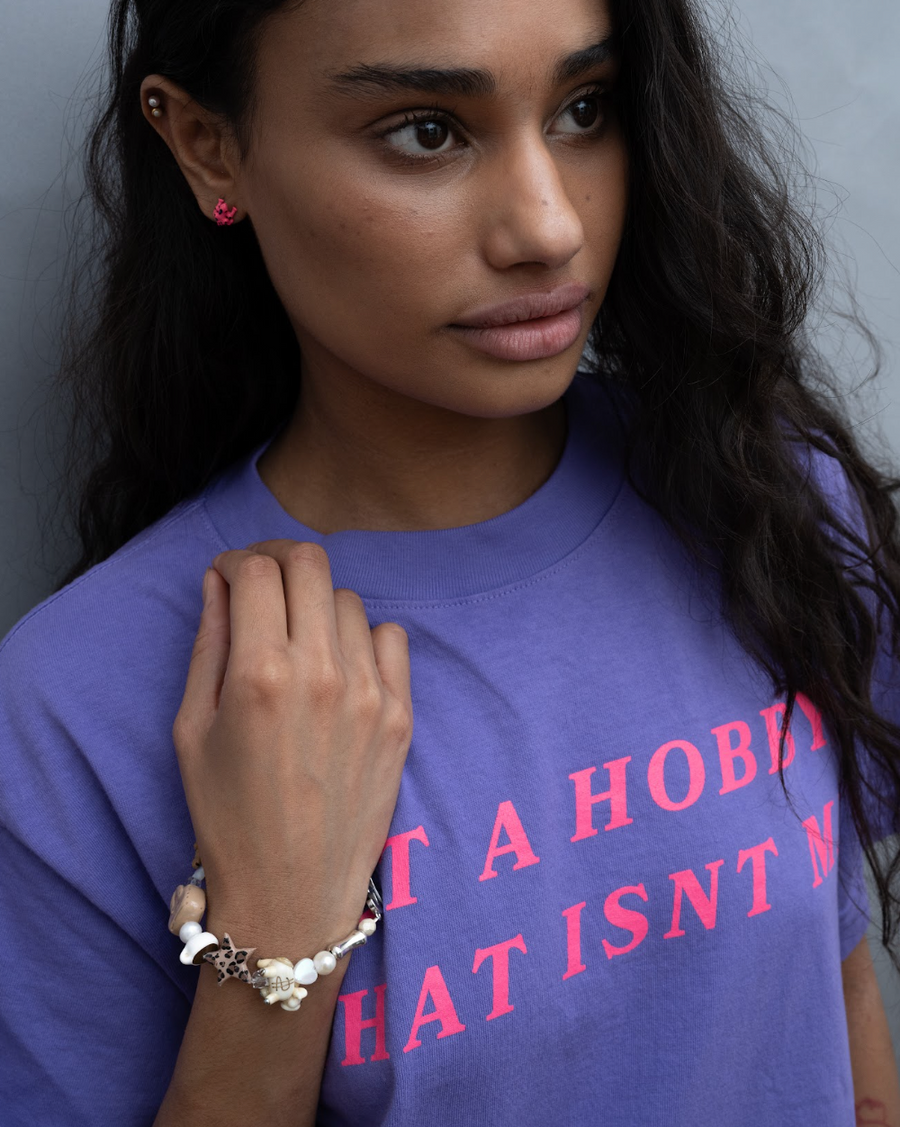 Woman in a purple slogan tee wearing a pink frog stud earring and the cream quiet lux bracelet