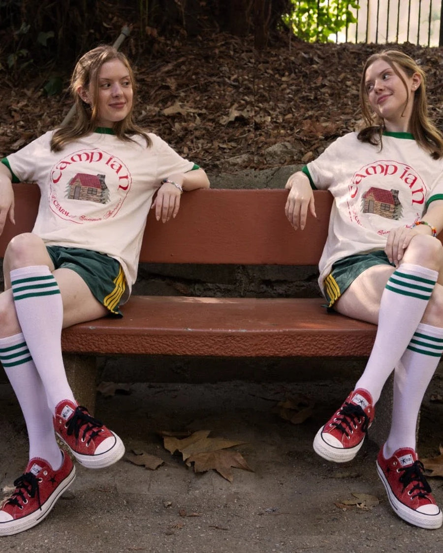 Two girls in tube socks and sneakers sitting on a bench wearing camp ian ringer tees