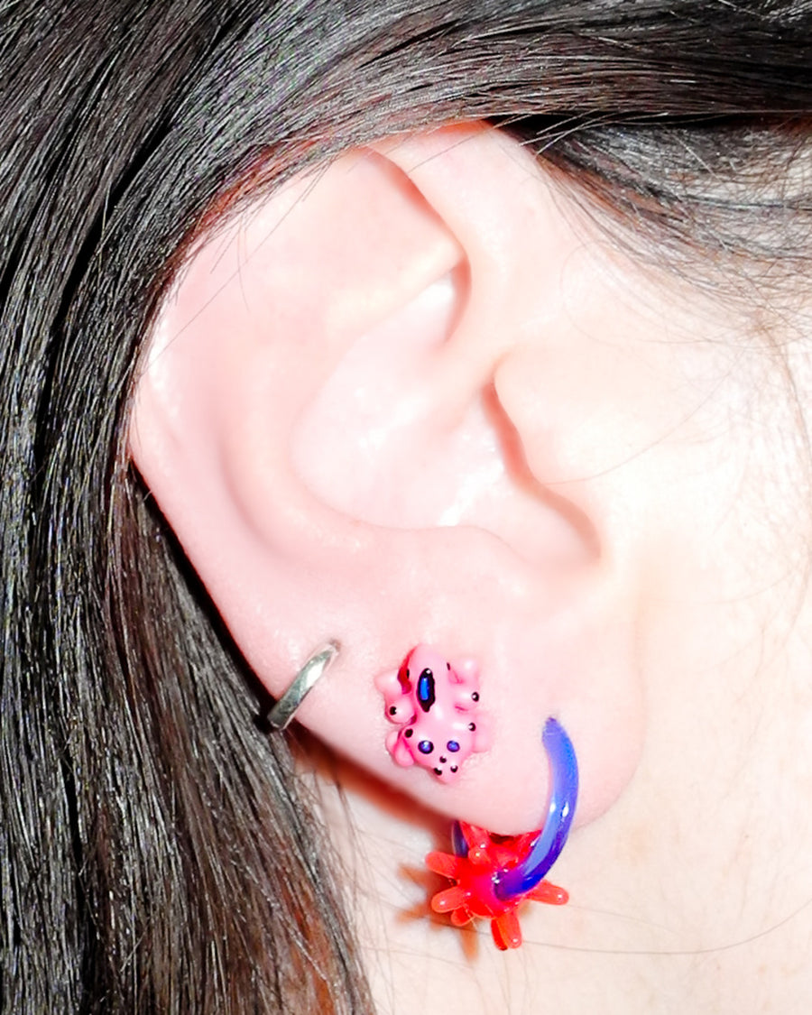 Close-up of an ear stacked with a pink frog stud, blue hoop and red spiky charm earring