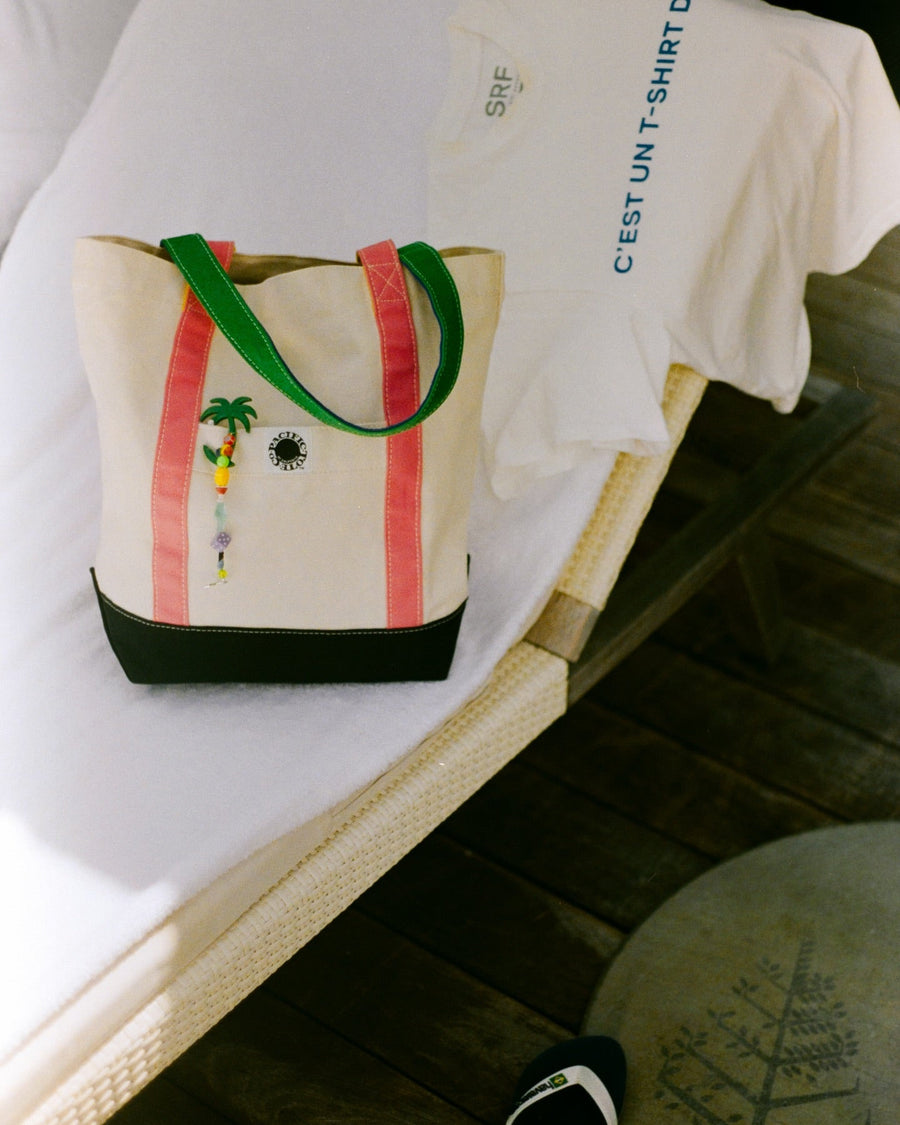 Canvas tote with the club mid palm tree beaded keychain resting on a lounge chair