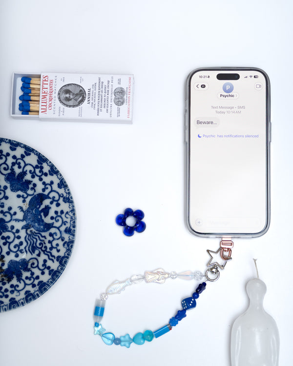 Blue and white beaded phone charm on a phone beside a blue willow plate and a blue flower – the lourdes phone charm