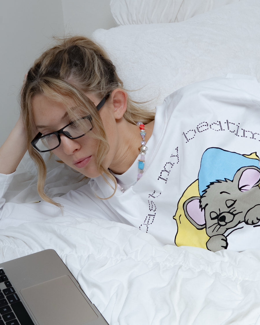 Woman in glasses lounging in bed with a laptop wearing the past my bedtime long sleeve tee