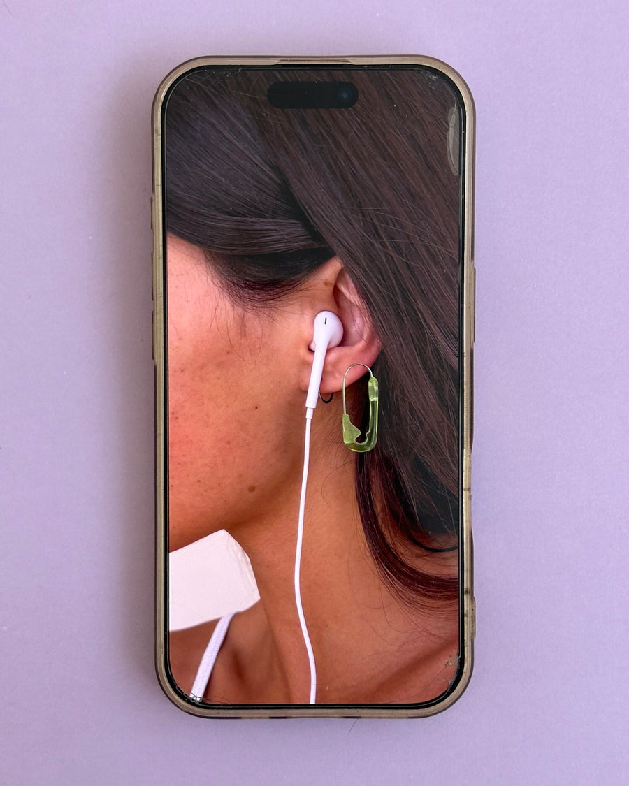 Phone screen on lilac showing an ear with a green safety pin earring and a white earbud